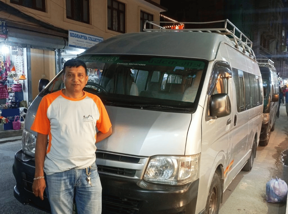 A white Hiace van parked on a roadside in Kathmandu, ready for the journey to Besisahar, with luggage loaded on the roof rack.